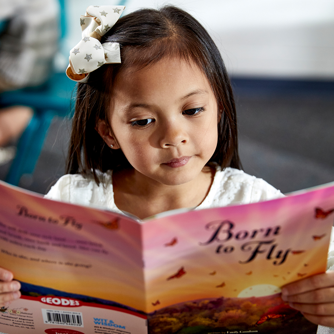 A young girl with a white bow in her hair sits in a classroom, reading a book titled Born to Fly. The book's cover features an illustrated sunset with butterflies and the title in bold, purple letters. The girl's expression is attentive and focused.