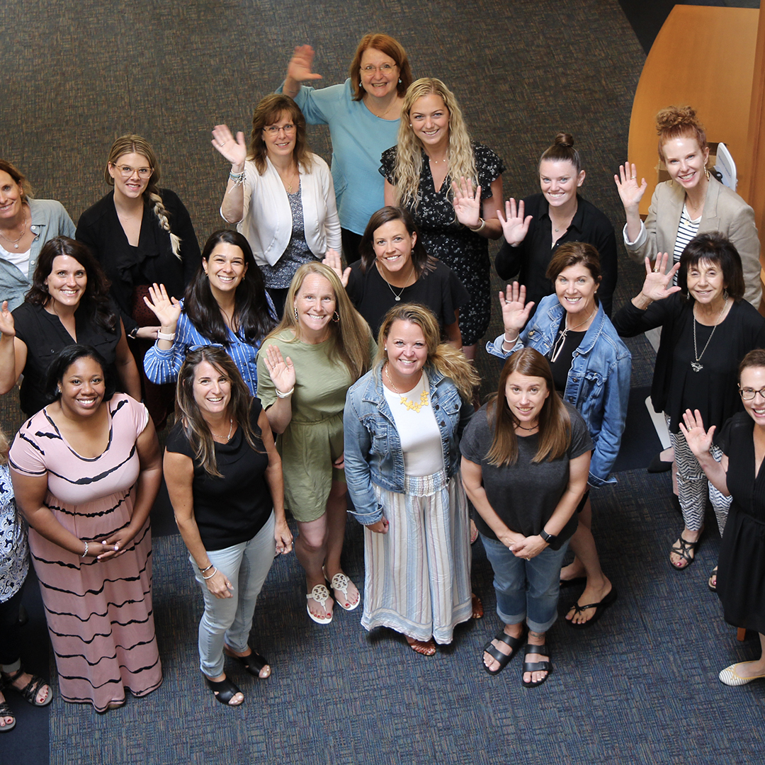 A group of 20 people, mostly women, stand indoors on a carpeted floor. They are smiling and waving at the camera. The group appears joyful and diverse, with varying styles of clothing and hair. The background includes a desk and a chair in the upper right corner.