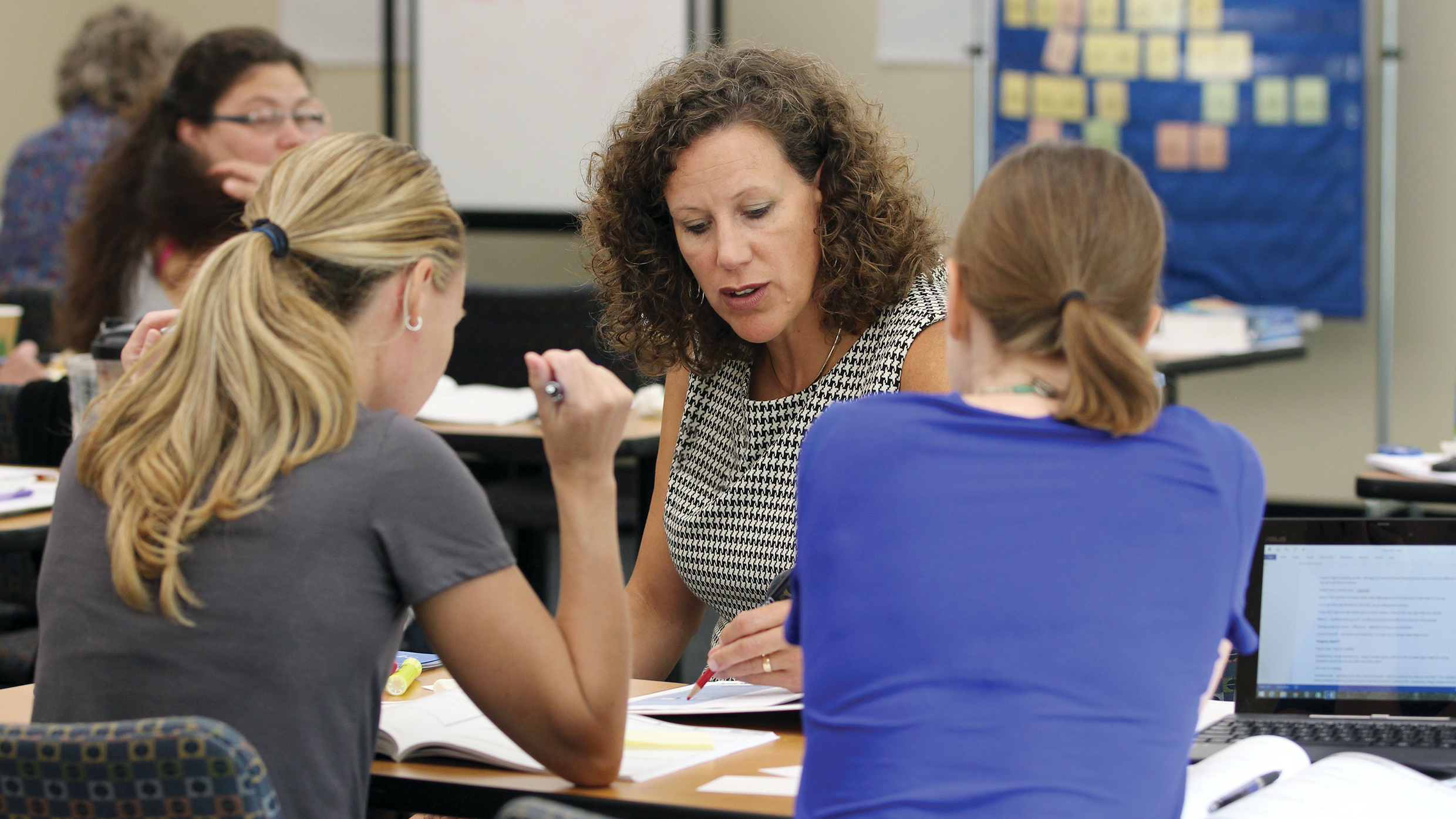 Three women sit at a table engaged in discussion, with open books and laptops in front of them. The woman in the middle, with curly hair, appears to be explaining something. In the background, other people are engaged in group activities, and a bulletin board is visible.
