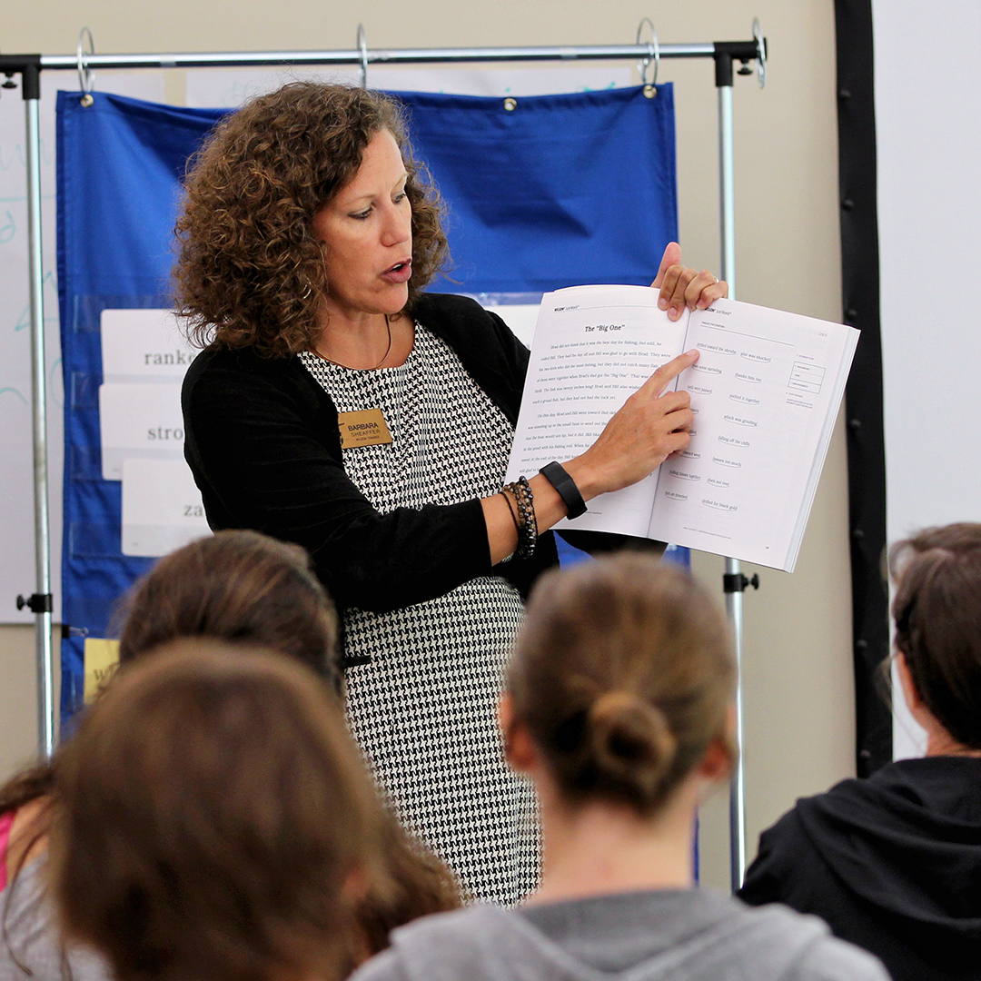 A woman with curly hair is standing and pointing to a page in a binder while speaking in front of a group of people. She is wearing a checkered dress and a name badge. The attendees are seated and focused on her presentation.