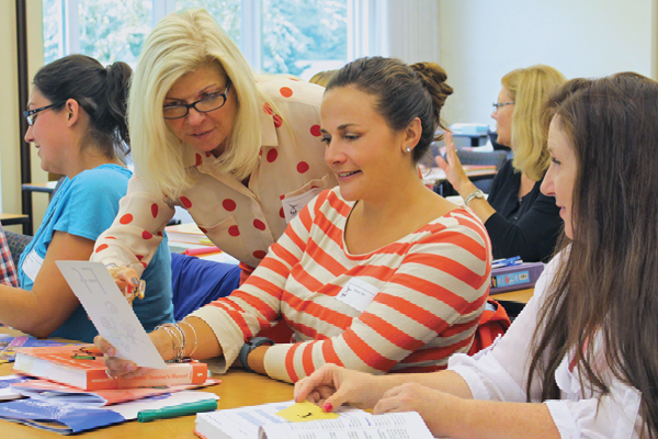 A group of women are sitting around a table, engaged in a discussion. One woman in a polka dot blouse is showing a paper to another woman in a striped shirt. Books and papers are scattered on the table, and a window is visible in the background.