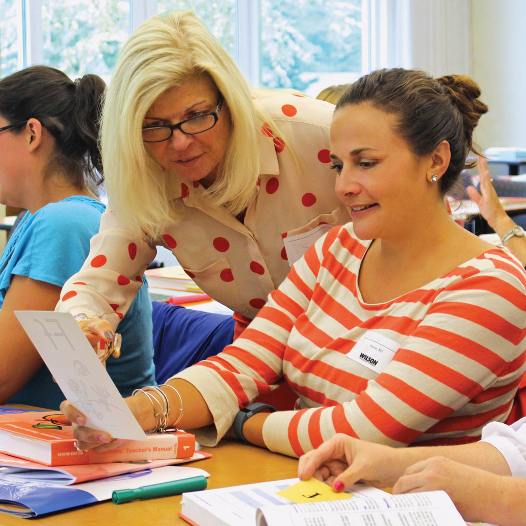 A woman with glasses and a polka dot shirt assists another woman with a worksheet. They are seated at a table with books and papers. Other people are visible in the background, engaged in similar activities.