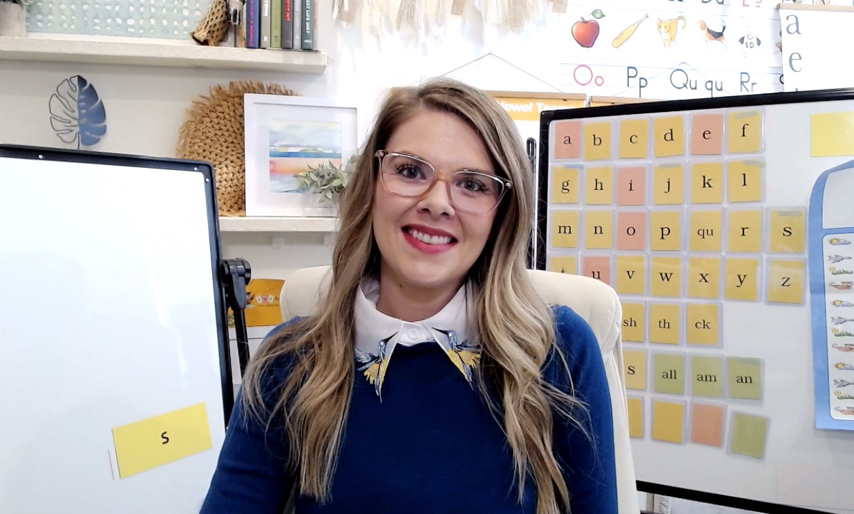 A woman with long hair and glasses is smiling while sitting in a classroom setting. Behind her are educational charts with letters and words, a whiteboard, and shelves with books and decorations.
