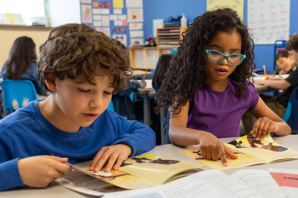 Two children are reading books at a classroom table. The boy on the left is wearing a blue shirt and has curly hair. The girl on the right, with curly hair and blue glasses, points to her book. Other students are seen studying in the background.