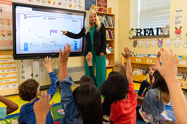 A teacher stands at the front of a classroom, pointing at a smartboard that displays the word "you." Several young students sit on a colorful carpet with their hands raised. The classroom is decorated with books, posters, and educational materials.