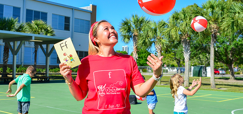 A woman in a red shirt holds a card with a letter F while smiling and tossing a beach ball outside. Children in the background play with beach balls on a sunny day. Palm trees and a building are visible.