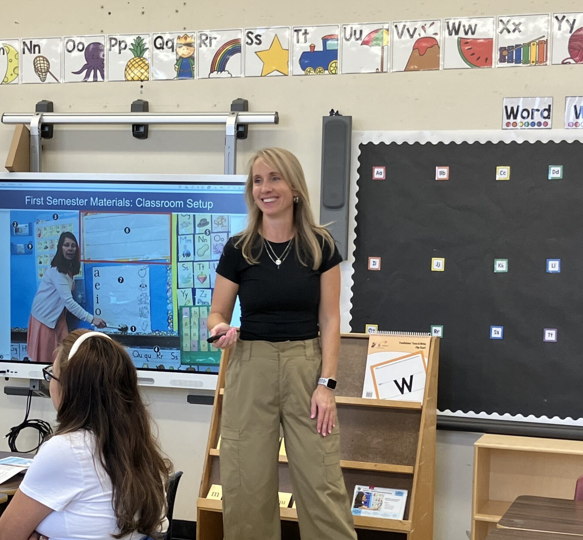 A teacher in a black shirt and tan pants stands in front of a classroom, smiling and gesturing. Behind her is a digital screen displaying First Semester Materials: Classroom Setup with a photo, and decorative borders on the walls.