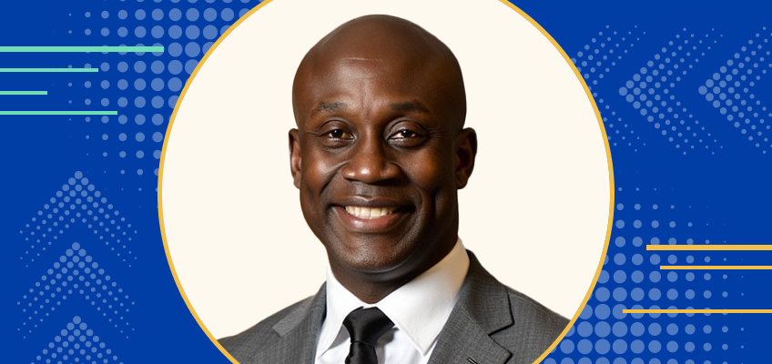 A person in a gray suit and black tie smiles against a white background. The image has a blue border with dotted patterns and yellow lines.