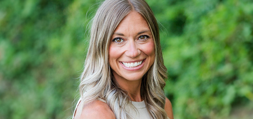 A woman with long, blonde hair smiles at the camera. She is wearing a sleeveless, light-colored top. The background is blurred with green foliage, suggesting an outdoor setting.