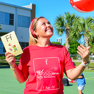 A smiling woman wearing a red shirt and headband catches a red balloon outdoors. She holds a card with the letter "F" printed on it. Palm trees and a building are in the background, and the scene suggests a fun, educational outdoor activity.