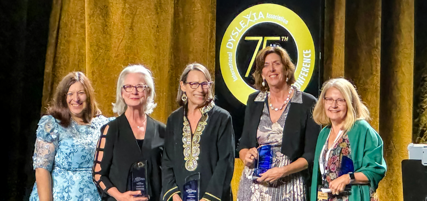 Five women stand together, smiling, each holding a blue award. They are in front of a gold curtain with a circular sign reading International Dyslexia Association 75th Conference.