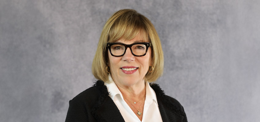 A person with shoulder-length blonde hair and glasses is smiling. They are wearing a black jacket over a white shirt. The background is a textured gray.