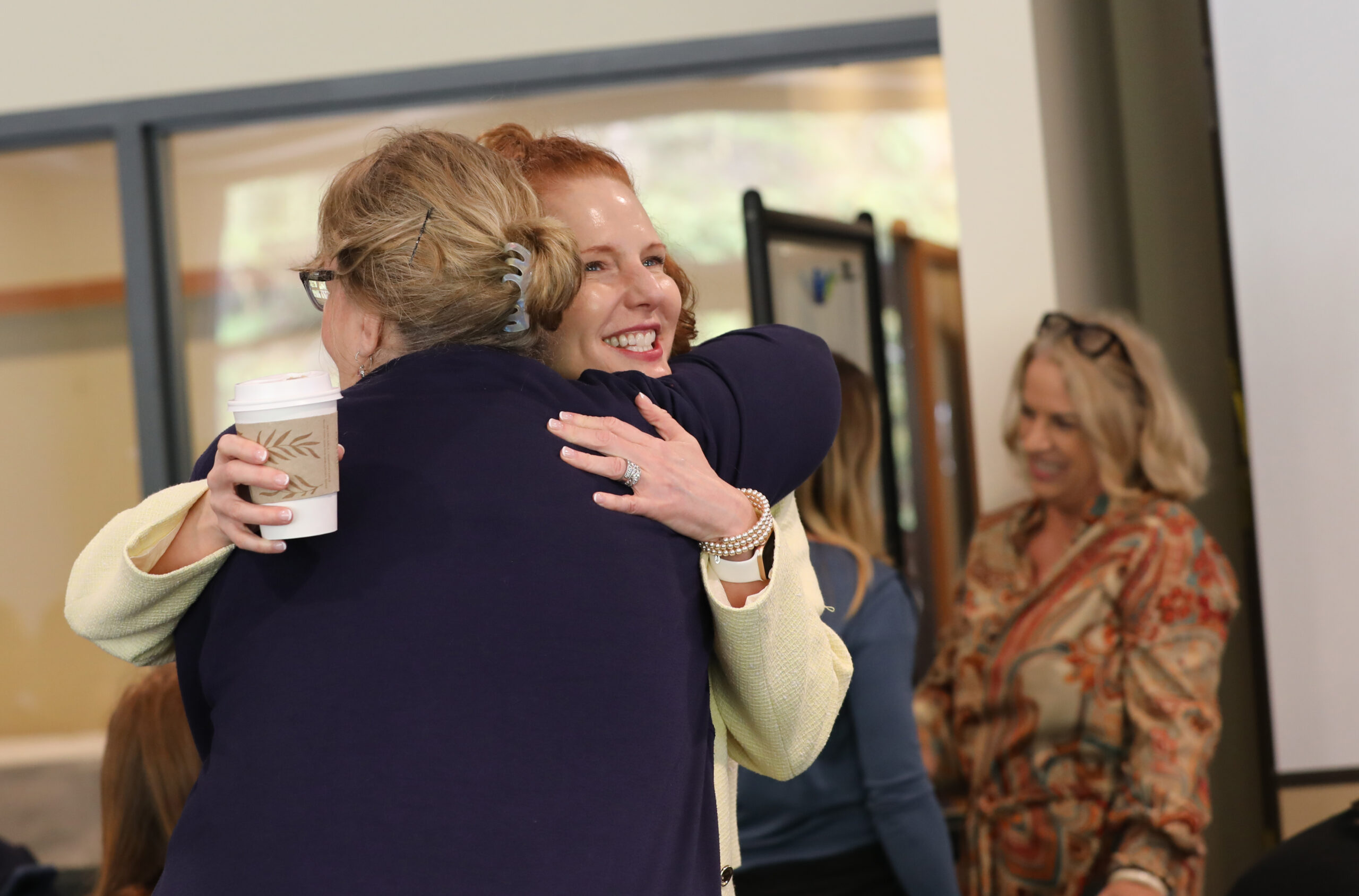 Two women share a warm embrace, one holding a coffee cup while the other smiles brightly. The woman facing the camera has red hair and is wearing a light yellow blazer, pearl bracelet, and a wedding ring. In the background, another woman with blonde hair, wearing glasses on her head and a patterned blouse, is engaged in conversation with someone off-camera. The setting appears to be a professional or community gathering.