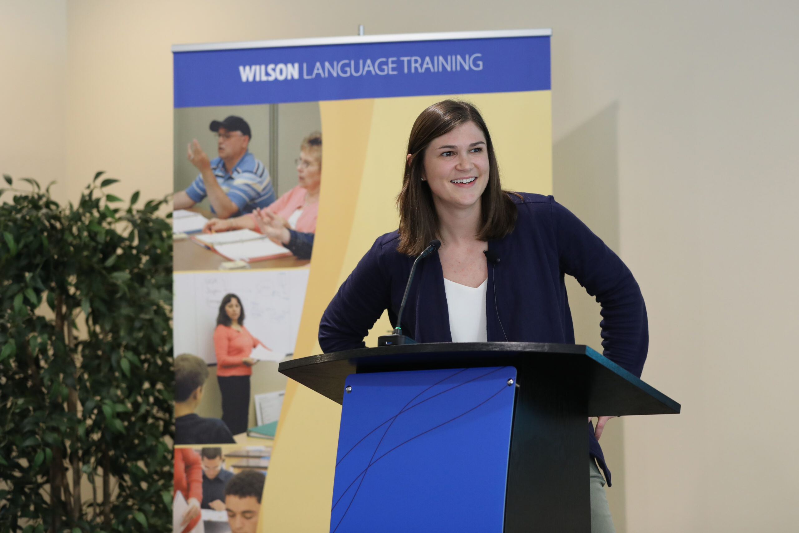 Keri Dixon, CEO of Wilson Language Training, stands behind a podium with a microphone, smiling as she speaks. She is wearing a navy blazer over a white top. Behind her is a large banner displaying the 'Wilson Language Training' logo along with images of people engaged in learning activities. A green plant is visible to the left.