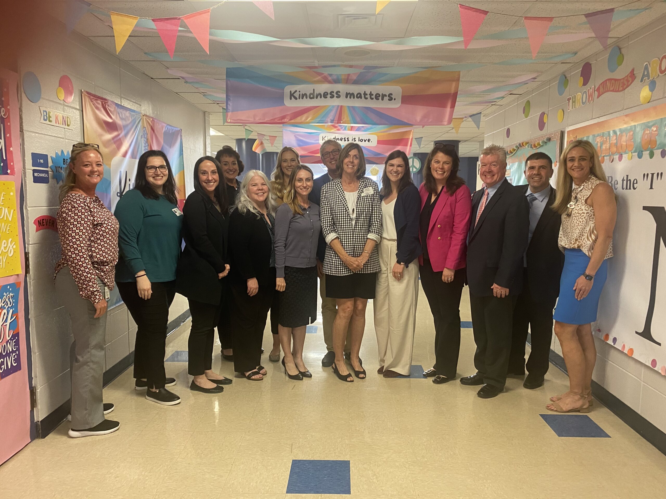 Barbara Wilson, Founder of Wilson Language Training, stands in the center of a group of professionally dressed people in a decorated school hallway. She is wearing a black and white checkered jacket and a black skirt. The hallway features colorful banners and posters with positive messages like 'Kindness matters' and 'Throw kindness around.' The group is smiling and posing for the photo.