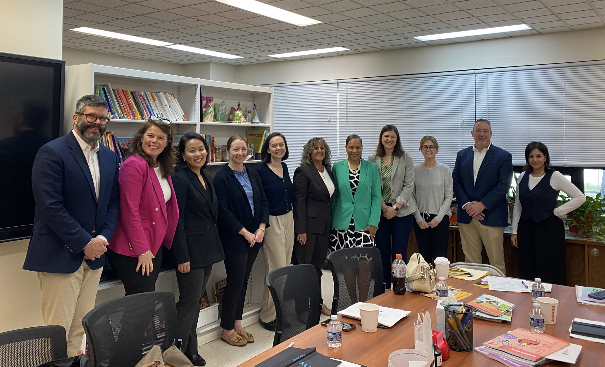 A group of eleven professionally dressed people stand together in a well-lit office or meeting room, smiling at the camera. Behind them are bookshelves filled with books and decorative items. A large conference table in the foreground is covered with notebooks, papers, water bottles, and office supplies.