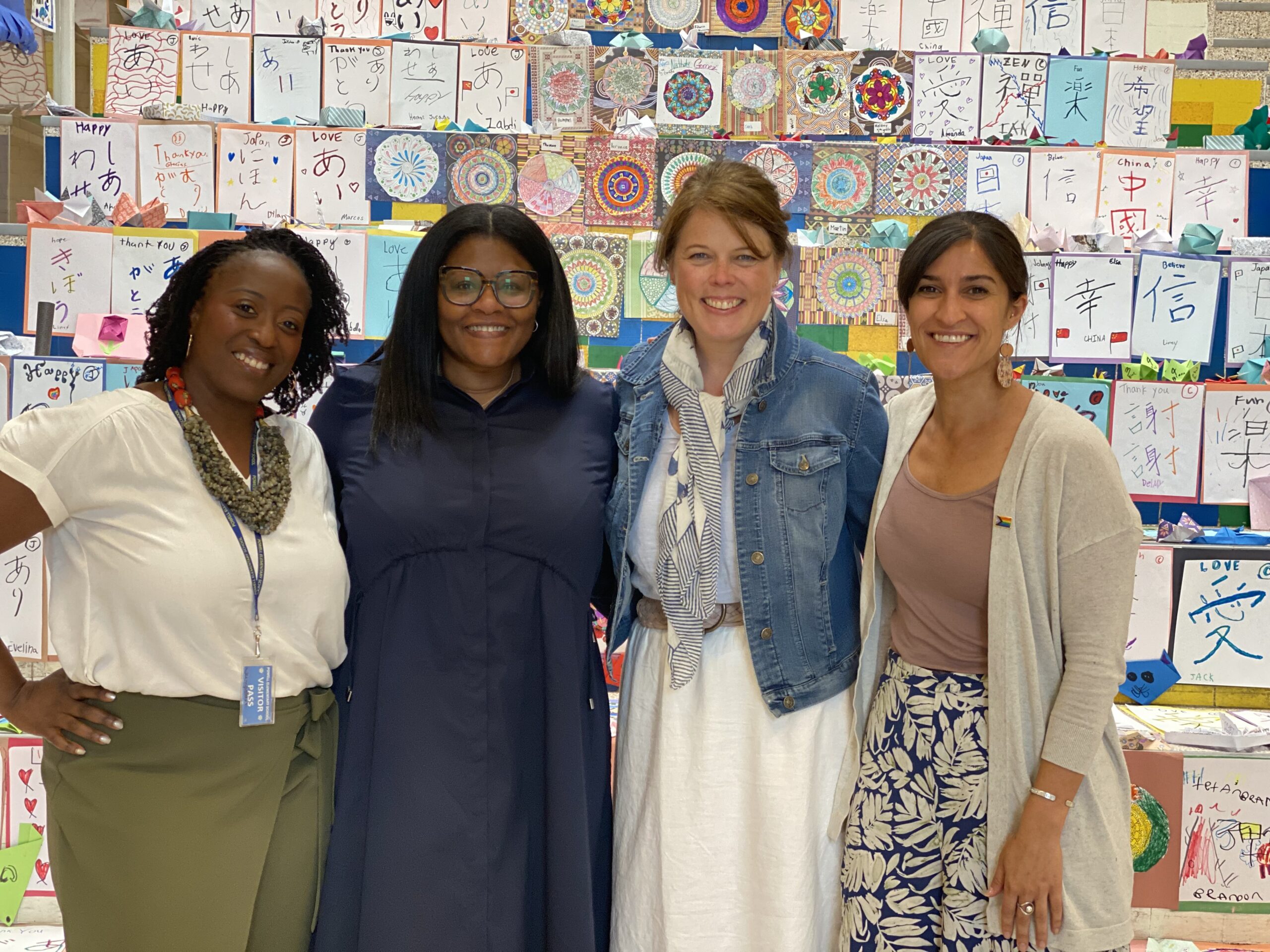 Four women stand together, smiling in front of a colorful display of student artwork featuring hand-drawn mandalas, Japanese characters, and messages of gratitude. They are dressed in a mix of casual and professional attire, including a white blouse with a statement necklace, a navy blue dress, a denim jacket over a white dress with a scarf, and a beige cardigan over a patterned skirt. The setting appears to be a school or community space, celebrating cultural diversity and creativity."