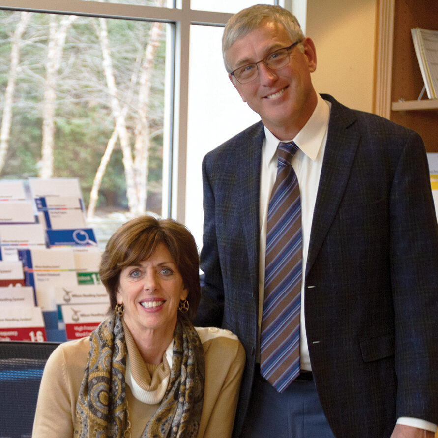 Barbara and Ed Wilson pose together in an office setting. Barbara, seated, wears a beige sweater with a patterned scarf and gold earrings, smiling warmly. Ed, standing beside her, wears a dark plaid blazer, white shirt, and striped tie, also smiling. Behind them is a window showing trees and a shelf with educational materials and books.