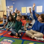 A diverse group of young students sit cross-legged on a colorful classroom rug, eagerly raising their hands. They are dressed in casual clothing and appear engaged, looking toward the teacher or lesson. The background features a bright blue wall decorated with student artwork, posters, and educational materials.