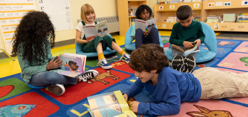 A group of five young children are reading books in a classroom setting. They sit and lie on a colorful alphabet rug featuring animal illustrations. The background includes bookshelves and educational posters, creating a warm, inviting reading environment.