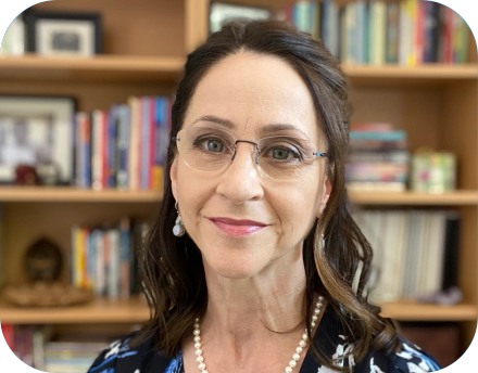 Bonnie Singer smiling in front of a bookshelf filled with colorful books and framed photos. She wears glasses, pearl earrings, a pearl necklace, and a floral-patterned top.