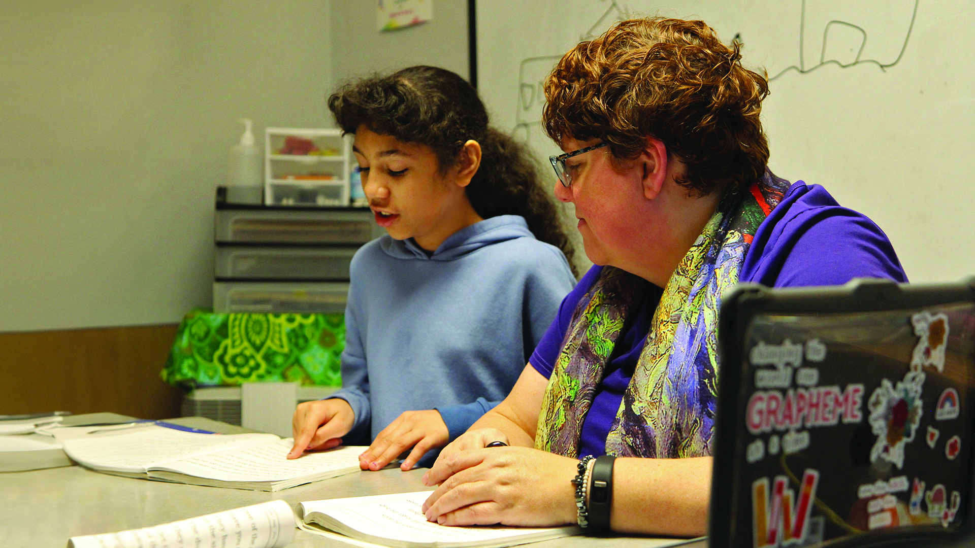 A student reading with her finger as their teacher looks on