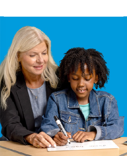 Adult woman and young girl seated at a table. The woman gently guides the girl's hand as she writes with a marker on a whiteboard.