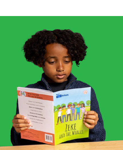 Young boy with curly hair reads a book titled "Zeke and the Whales" while seated at a desk.
