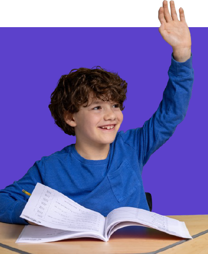 Smiling boy in a blue shirt raises his hand while looking to the side, seated at a desk with an open workbook.