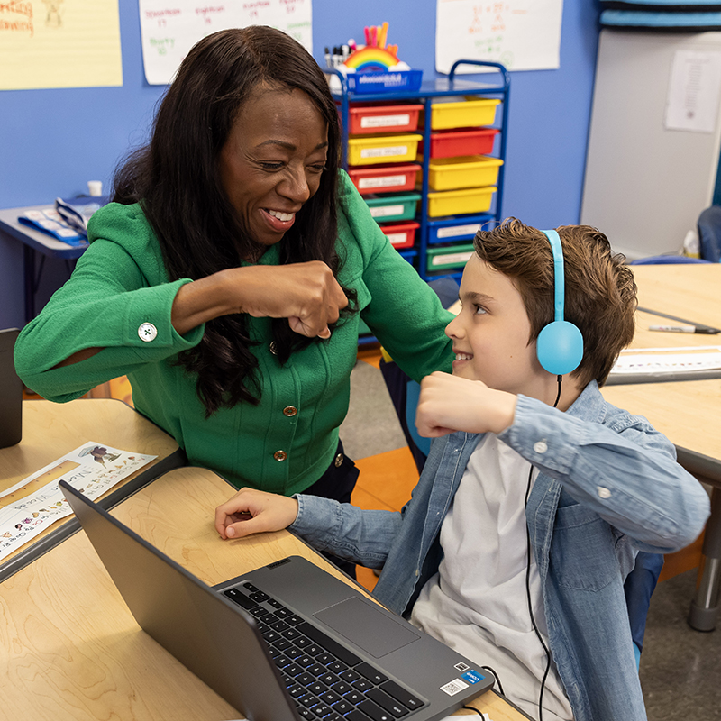 Teacher fist-bumping student with headphones at a laptop in a classroom.