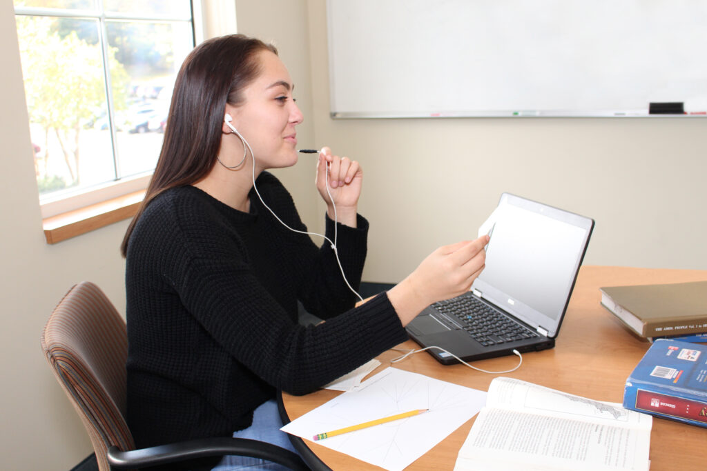 Young woman in black sweater with earbuds studies at desk with laptop