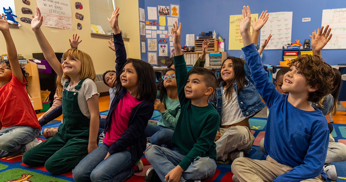 Diverse elementary students raising hands in a classroom, eager to learn.