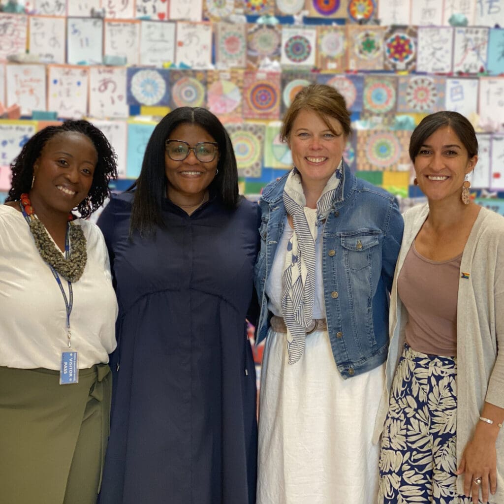 Four women smile in front of a colorful, art-covered wall.