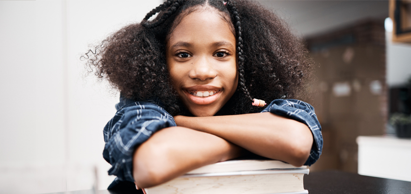 Young Black girl with braided hair smiling, resting arms on a stack of books.