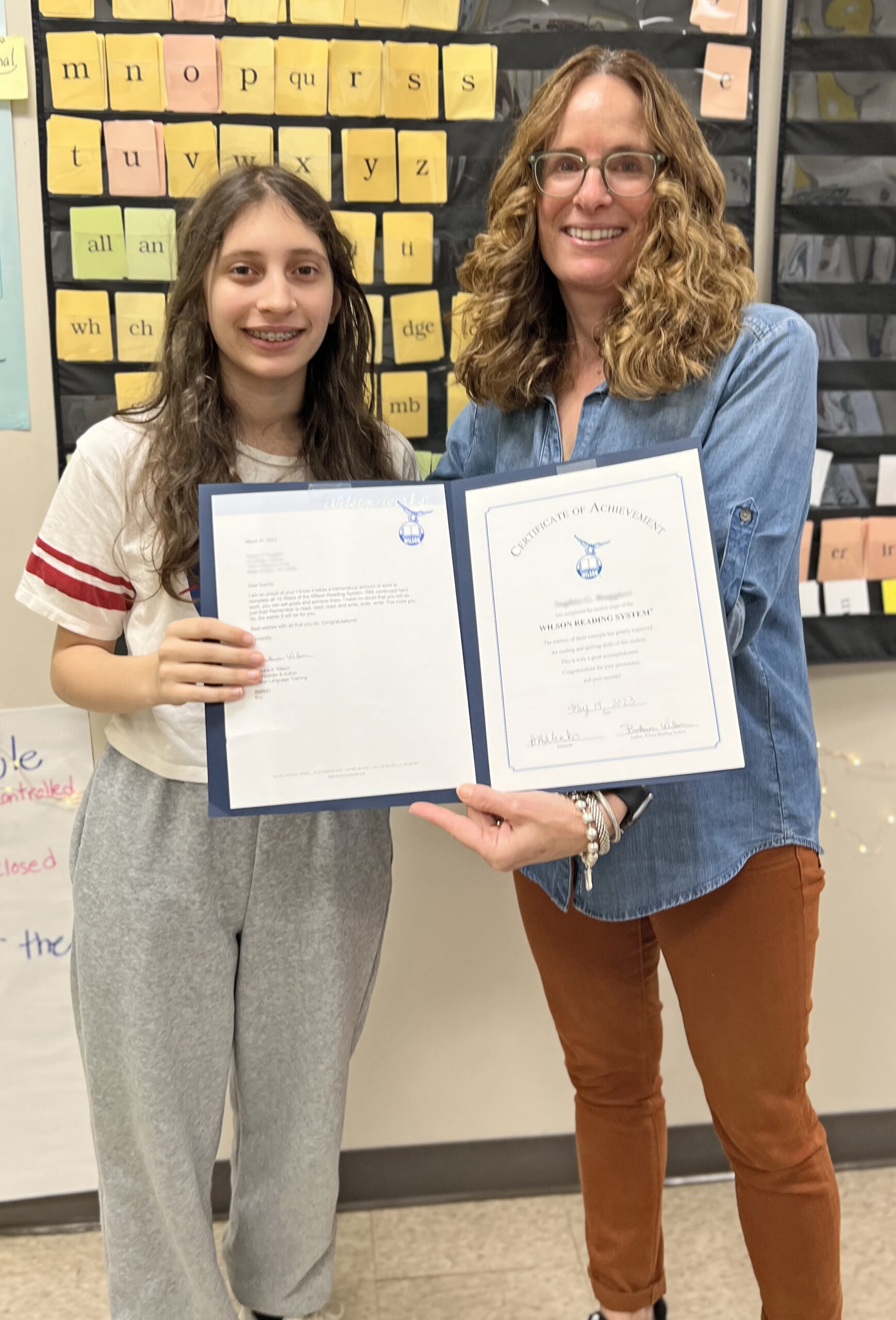 A student and teacher smiling while holding up a Certificate of Achievement together in a classroom. The certificate is displayed in a blue folder, and the background features an educational word wall with yellow and black tiles.